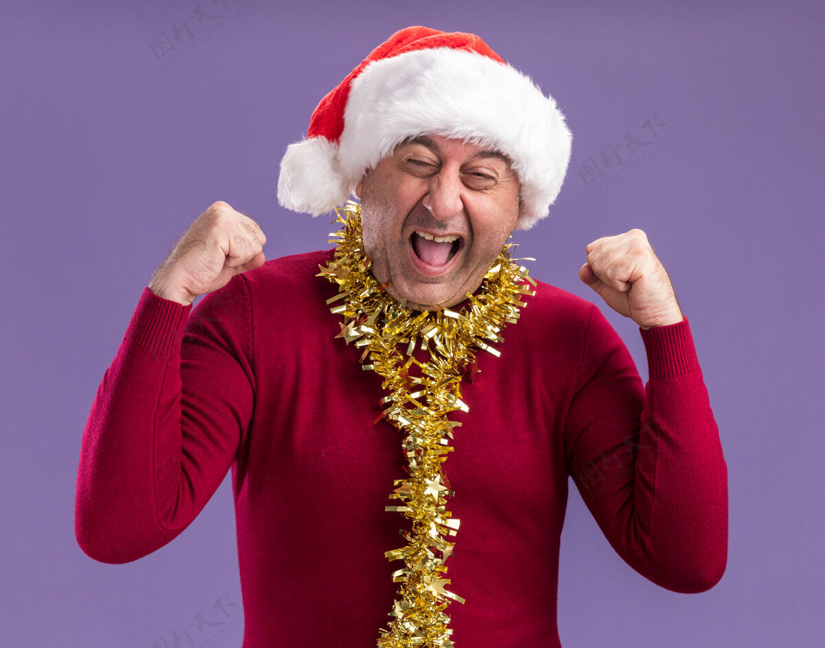 帽子中年男子戴着圣诞老人帽子 脖子上戴着金属丝 高兴而兴奋地握紧拳头尖叫着站在紫色背景上周围兴奋金属片