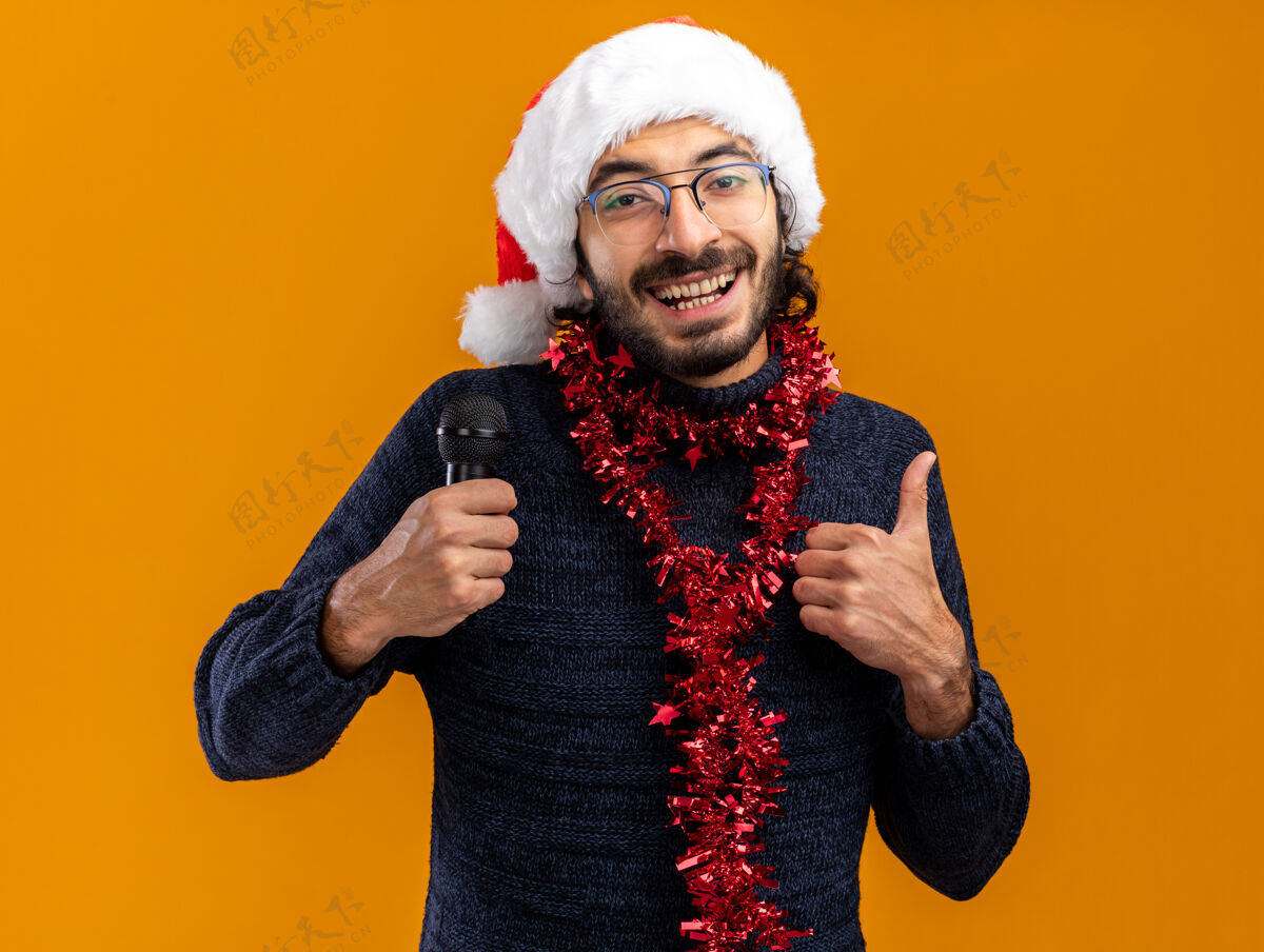 年轻微笑的年轻帅哥戴着圣诞帽 脖子上戴着花环 拿着麦克风 在橙色背景上孤立地竖起大拇指家伙显示微笑