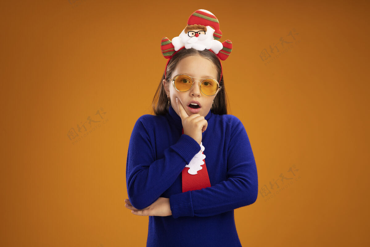高领毛衣小女孩穿着蓝色高领毛衣 打着红色领带 头上戴着有趣的圣诞戒指 站在橙色的背景下 惊讶地看着摄像机领带有趣圣诞节