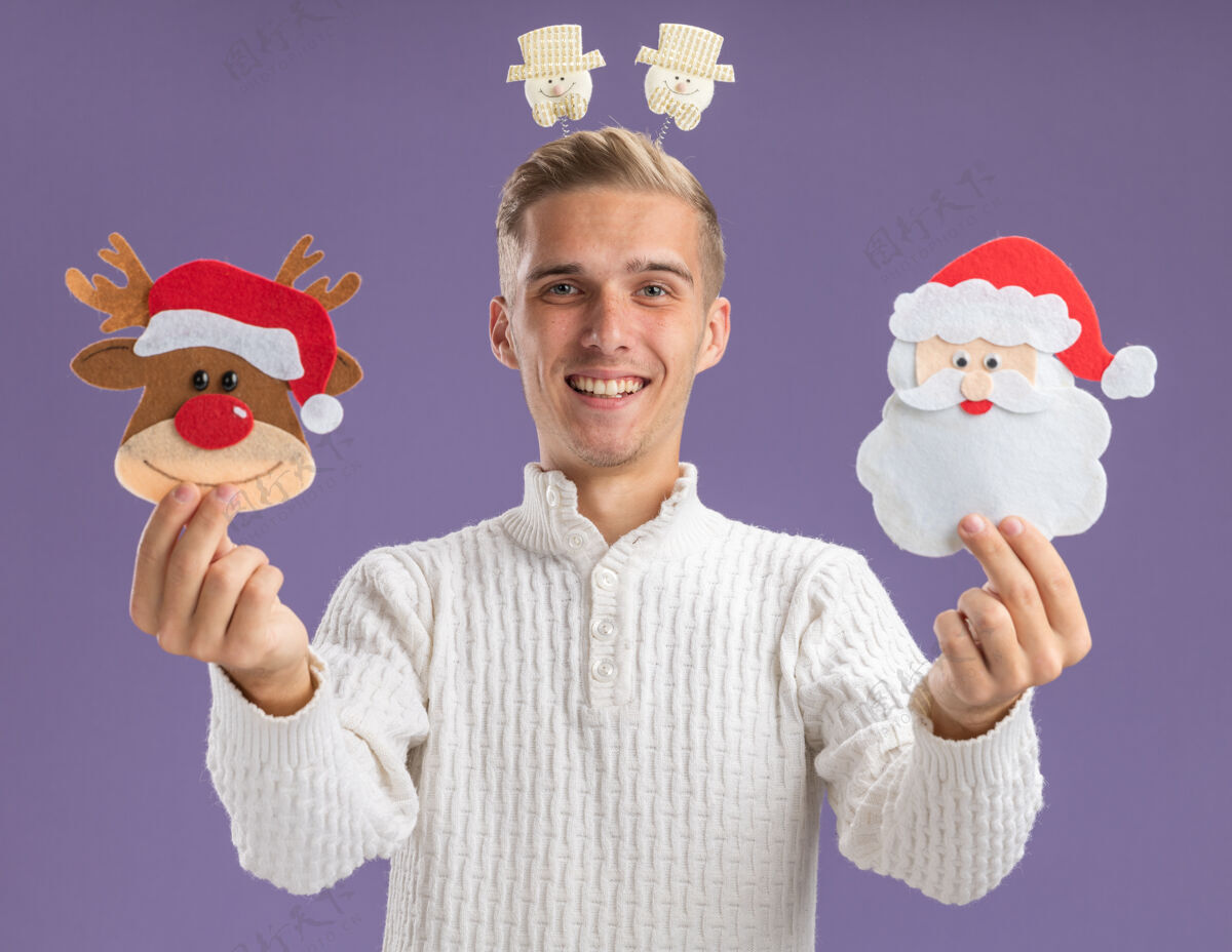圣诞节快乐的年轻帅哥戴着雪人头巾 向孤零零的紫墙上伸出圣诞纸饰品快乐装饰品雪人