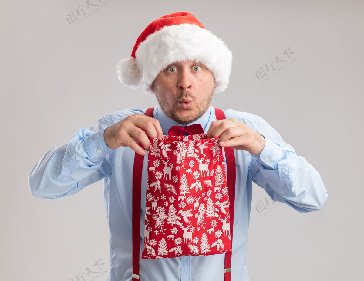 包年轻人戴着吊带领结 戴着圣诞帽 手里拿着装满礼物的红包 站在白色背景下惊讶地看着镜头帽子圣诞老人站着