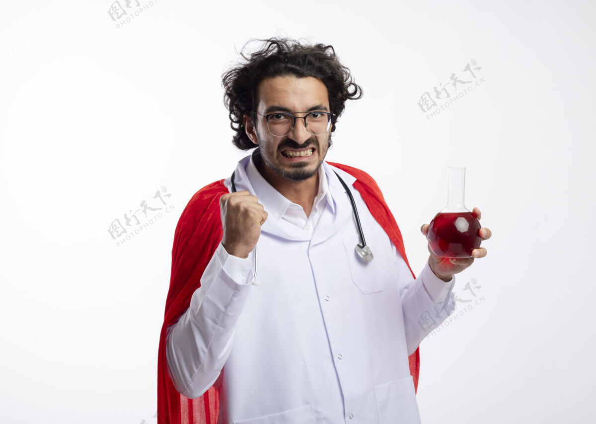 年轻人戴着眼镜 身穿医生制服 披着红色斗篷 脖子上戴着听诊器的未出狱的白人年轻人握紧拳头 把红色化学液体装在白墙上的玻璃瓶中男人化学品医生