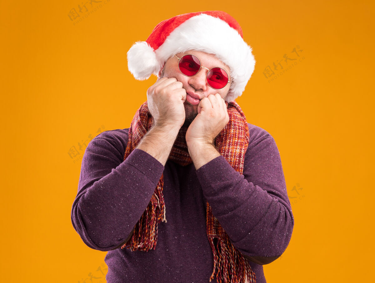 手无聊的中年男子戴着圣诞帽 脖子上戴着金箔花环 戴着眼镜 一边看着一边 手放在下巴上 橙色的背景上孤立着侧面无聊脖子