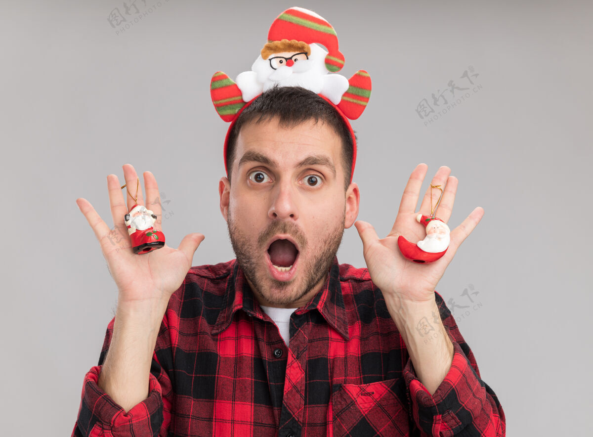 男人令人印象深刻的年轻白种人戴着圣诞老人头带拿着圣诞老人圣诞饰品隔离在白色的墙壁上印象深刻圣诞老人年轻人