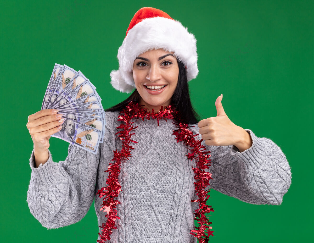 年轻快乐的年轻白人女孩戴着圣诞帽 脖子上戴着金箔花环 手里拿着钱 在绿色的墙上孤立地竖起大拇指周围快乐圣诞节