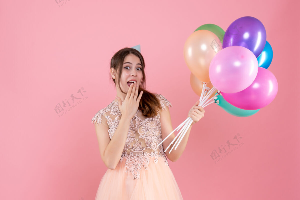 漂亮快乐的派对女孩 戴着派对帽 手里拿着气球 把她的手放在粉色的嘴巴上公主气球帽子