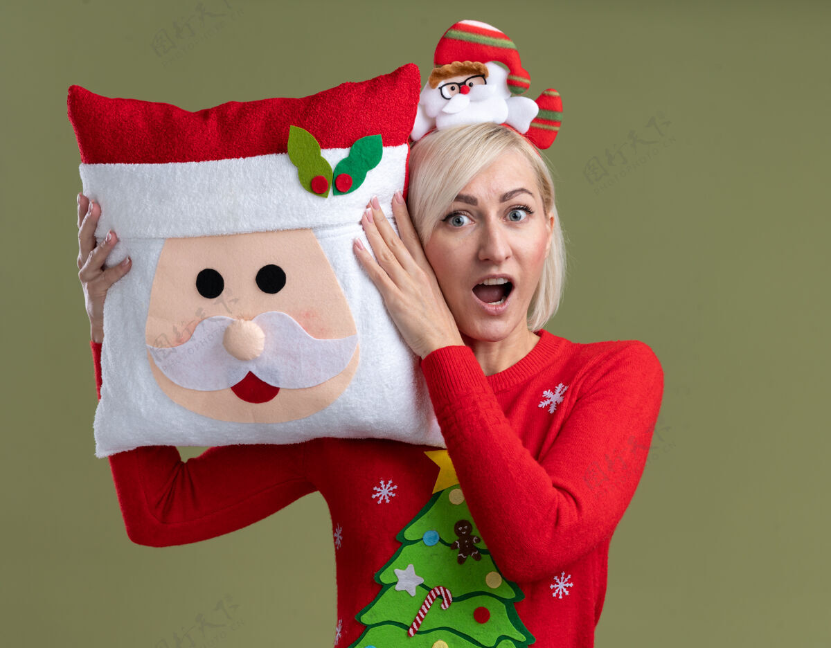 圣诞老人令人印象深刻的中年金发女人戴着圣诞老人的头带和圣诞毛衣抱着圣诞老人的枕头触摸它孤立的橄榄绿墙上的头橄榄毛衣头