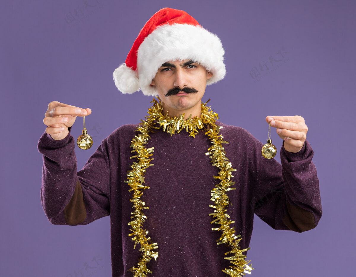 站着一个留着胡子的年轻人戴着圣诞老人帽子 脖子上戴着金属丝 手里拿着圣诞球 严肃的脸站在紫色的背景上看着摄像机举行脖子脸