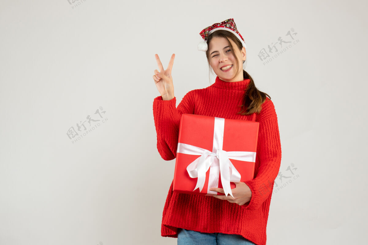 礼物戴着圣诞帽的快乐女孩拿着礼物站在白色的墙上做胜利的标志女孩成人快乐