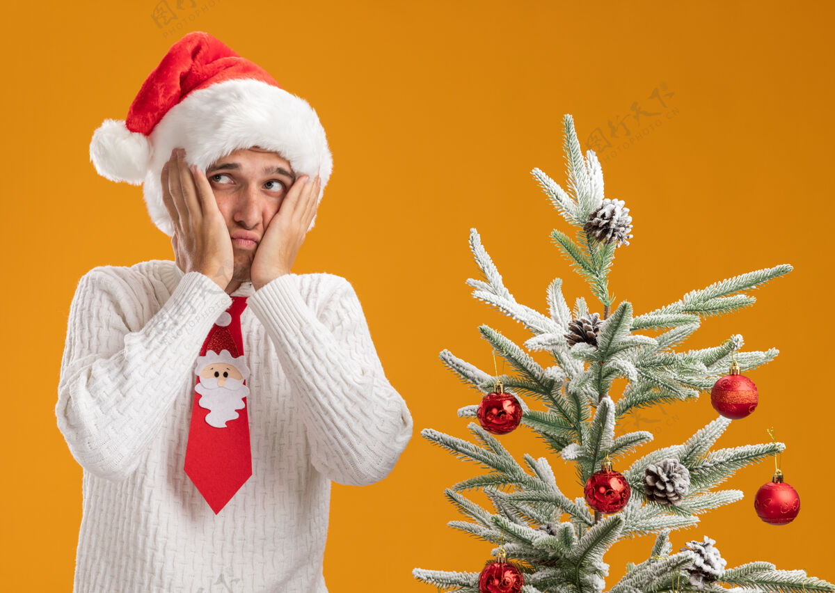 小伙子戴着圣诞帽 系着圣诞老人领带的年轻帅哥站在装饰好的圣诞树旁 手放在脸上看着橙色背景上孤立的一面帅气圣诞老人脸