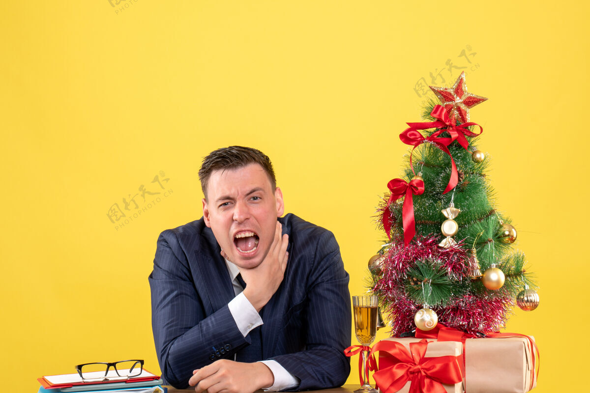 商人前视图愤怒的男子勒死自己的手坐在桌旁圣诞树和礼物上的黄色墙壁桌子生意坐着