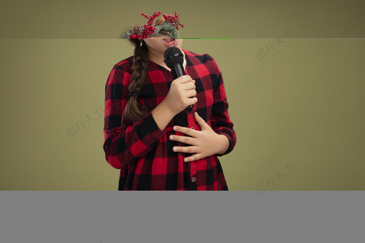 站着快乐和积极的小女孩戴着圣诞花环在格子裙手持麦克风唱歌站在绿色的背景衣服唱歌检查