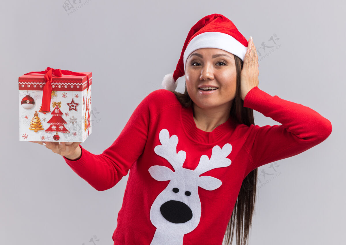 礼物快乐的年轻亚洲女孩戴着圣诞帽 拿着毛衣 手放在脸颊上 背景是白色的放着毛衣拿着