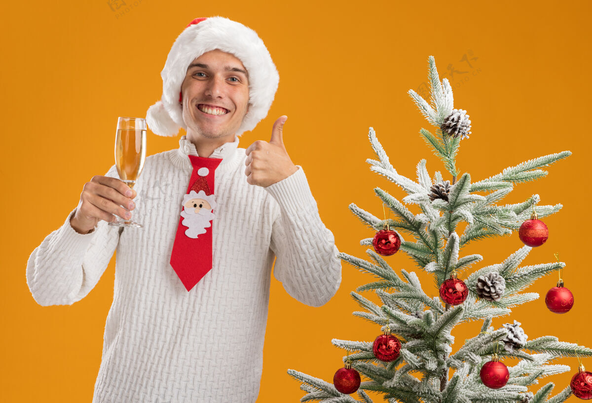快乐快乐的年轻帅哥戴着圣诞帽 打着圣诞老人的领带 站在装饰好的圣诞树旁 手里拿着一杯香槟 竖起大拇指孤立地站在橙色的墙上圣诞老人玻璃杯领带