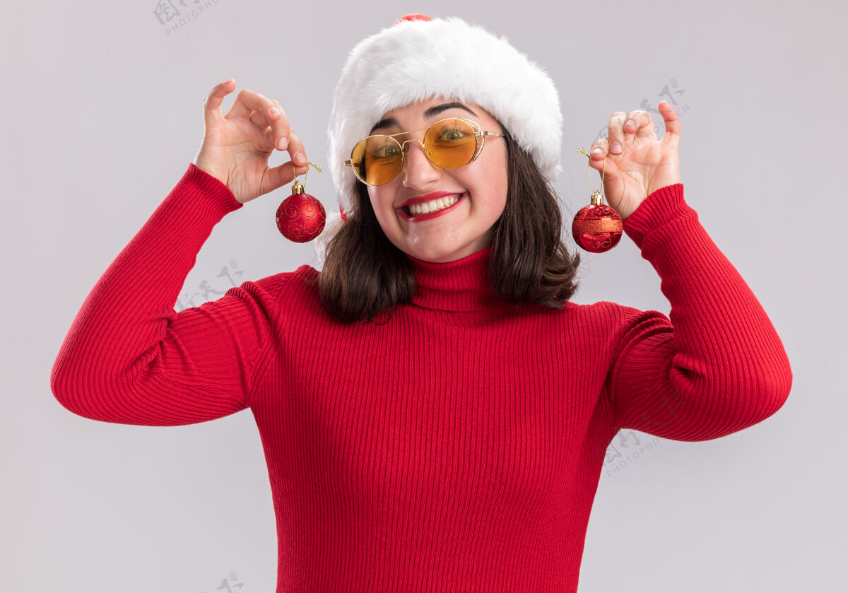 举行穿着红色毛衣 戴着圣诞帽 戴着眼镜 拿着圣诞球 站在白色背景下 微笑着看着镜头的快乐女孩圣诞节广泛站着