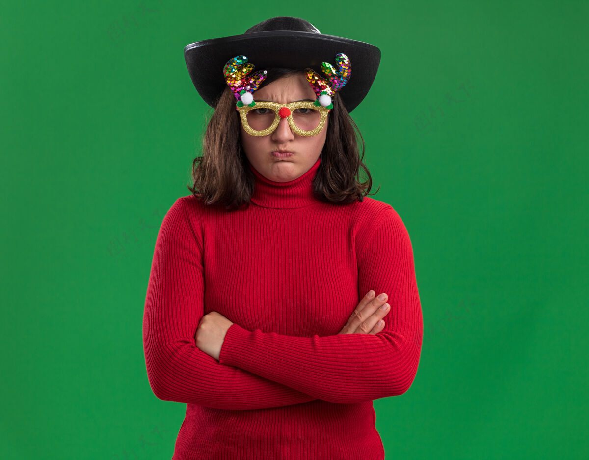 毛衣穿着红毛衣的小女孩戴着滑稽的眼镜和黑帽子 不高兴地交叉双臂站在绿色的背景上看镜头年轻胳膊不高兴
