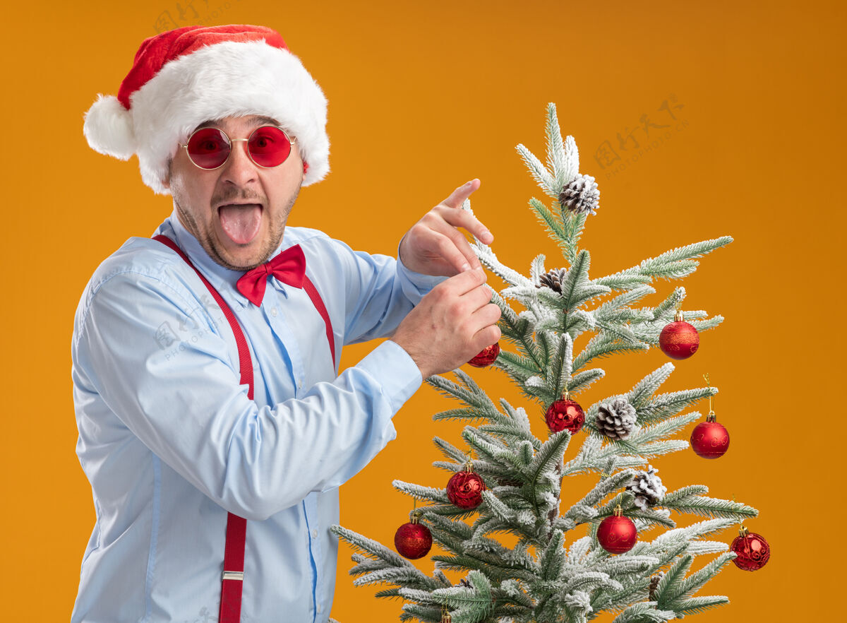 年轻快乐的年轻人戴着吊带领结 戴着圣诞帽 戴着红眼镜 站在圣诞树旁 把玩具挂在树上 高兴又惊讶地伸出舌头 遮住橙色的背景圣诞树帽子惊讶