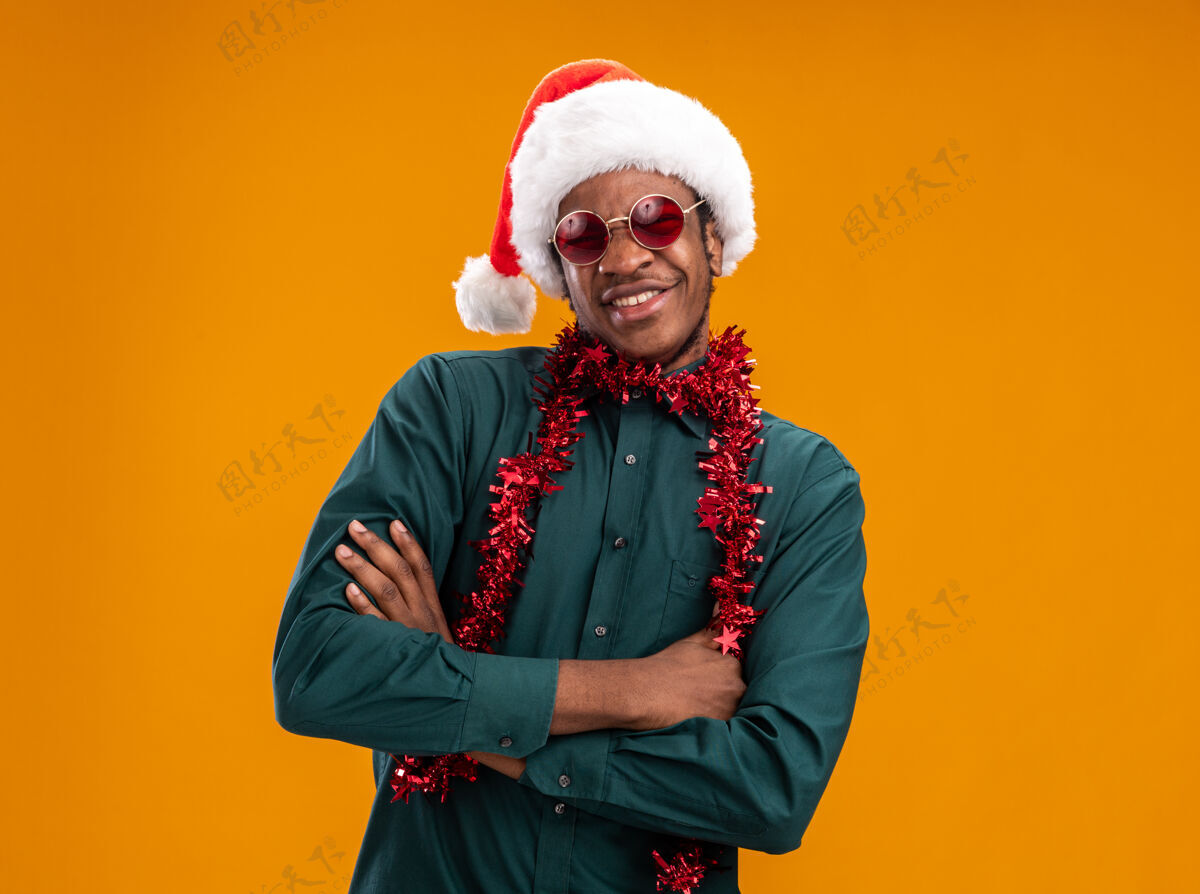微笑快乐的非洲裔美国人戴着圣诞帽 戴着花环 戴着太阳镜 面带微笑 双臂交叉 站在橙色背景上看镜头手臂花环非洲
