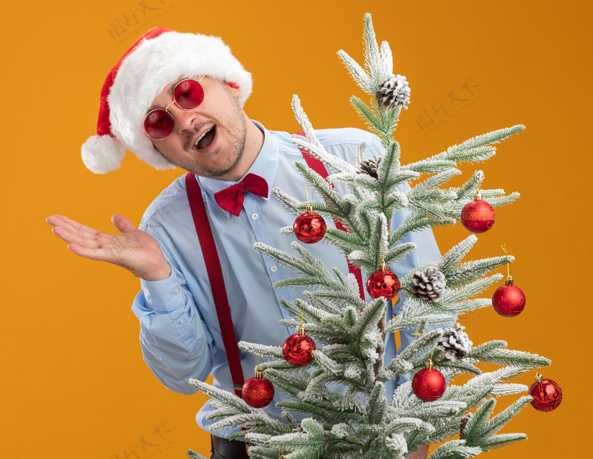 心情年轻人戴着吊带领结 戴着圣诞帽 戴着红眼镜 站在橙色背景下的圣诞树旁 兴高采烈地看着镜头好圣诞树站着