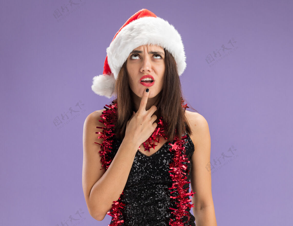 脖子迷茫的抬头年轻漂亮的女孩戴着圣诞帽 脖子上戴着花环 手指放在下巴上 紫色背景上孤立着女孩帽子手指