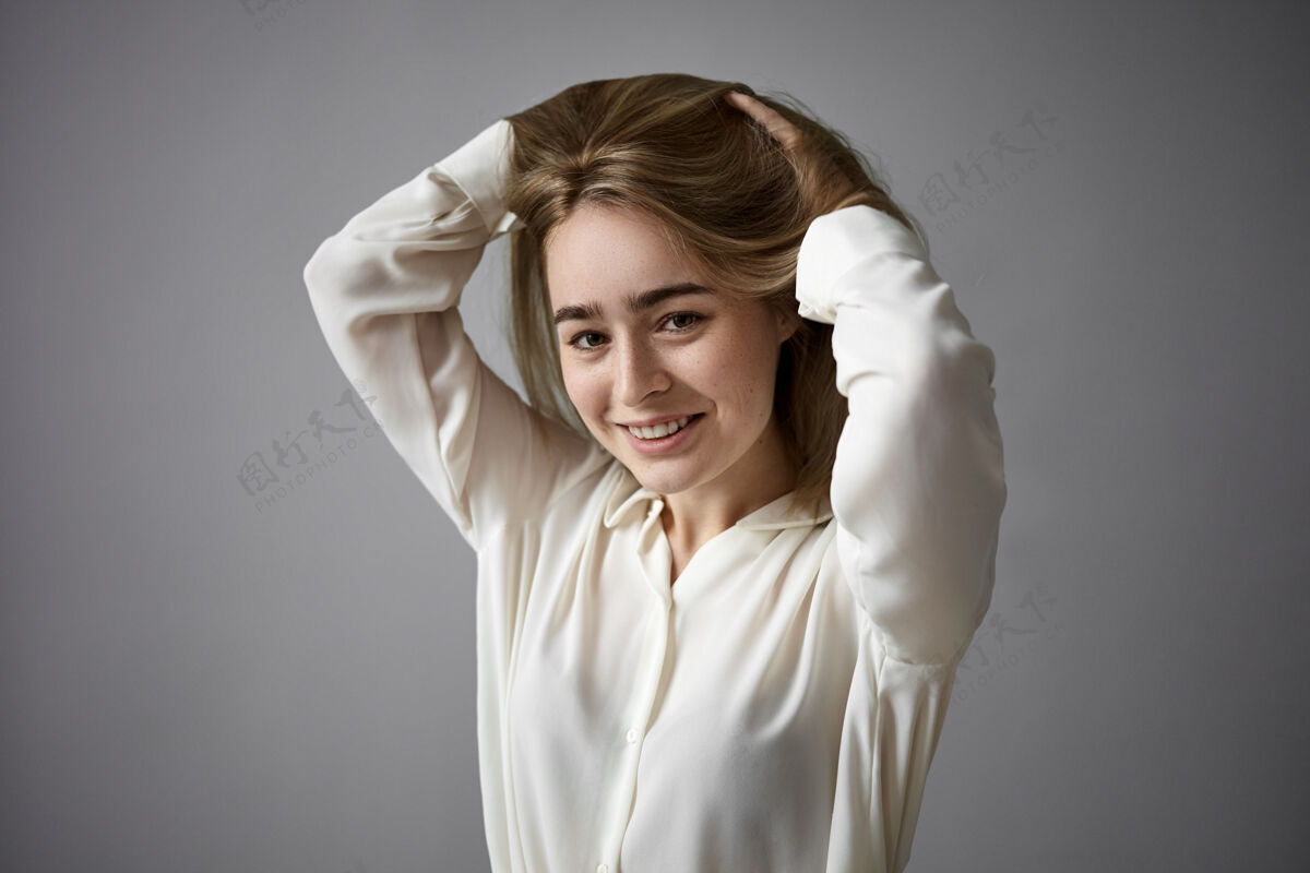 衬衫正面开朗的年轻女子穿着优雅的白色上衣 在镜头前愉快地微笑 抚摸着她的长发 为新发型而欢欣鼓舞人 时尚和风格的概念自信积极脸