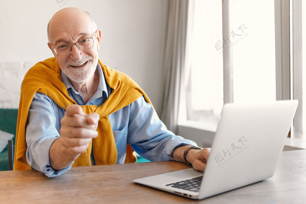 胡须帅气的 快乐的 兴奋的 留着灰胡茬的老男人 坐在现代办公室里的笔记本电脑上 坐在靠窗的办公桌旁 微笑着 用手指着相机选择性聚焦欢快商人休闲