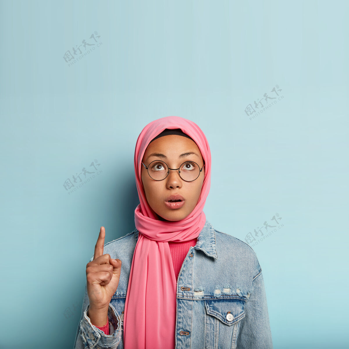 穆斯林惊讶的年轻穆斯林女子用一个食指指着 专注向上 有着关切的表情 蒙着头促销指向反应