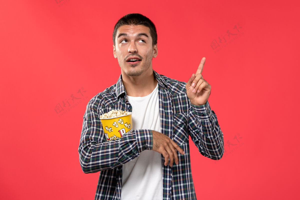 男孩正面图年轻男子手持爆米花包 在浅红墙电影院摆姿势年轻男性乐器观点