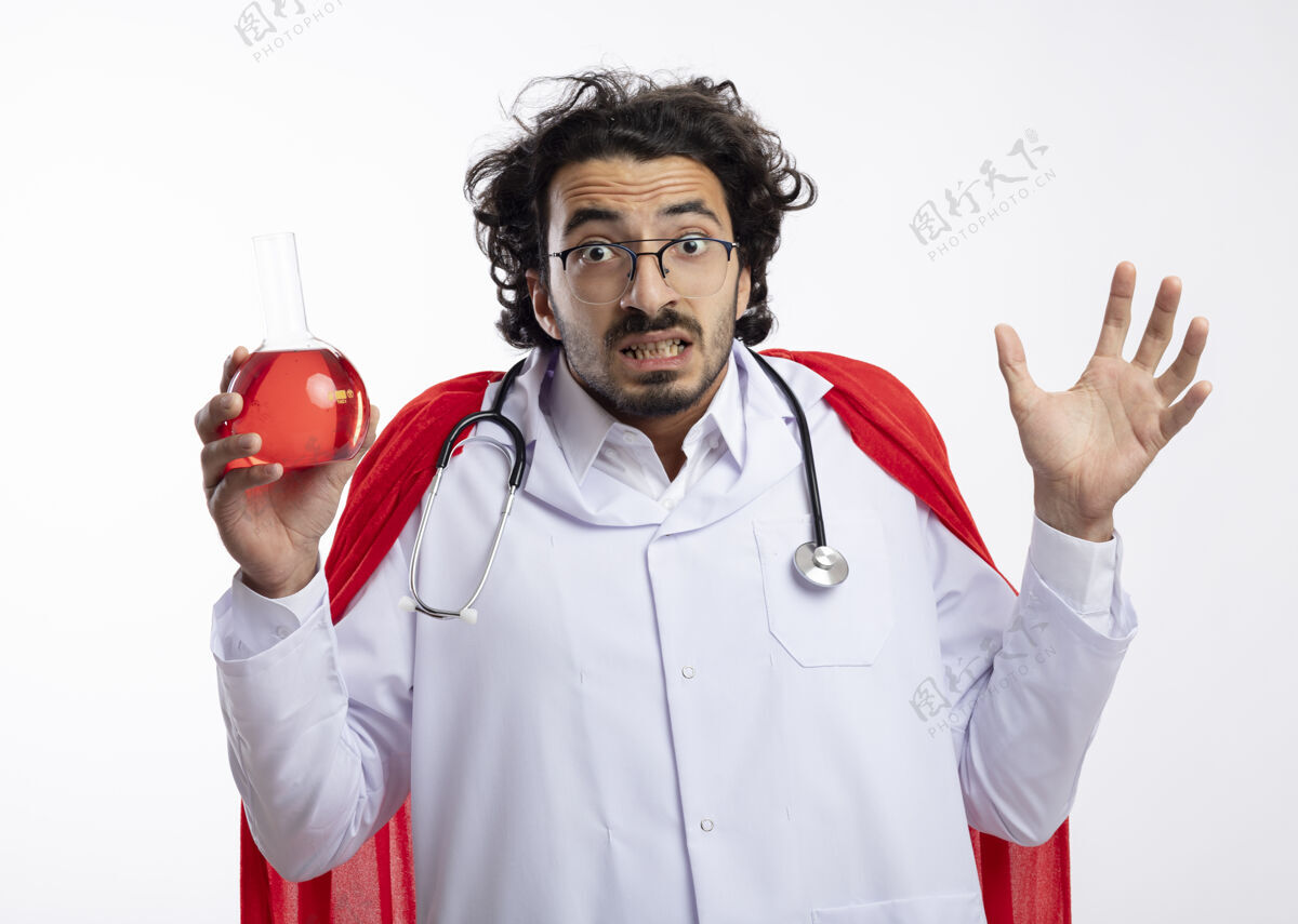 戴着焦急的白人年轻人戴着眼镜 身穿医生制服 披着红色斗篷 脖子上戴着听诊器 举起手站在玻璃瓶中 手里拿着红色的化学液体抬起年轻人男人