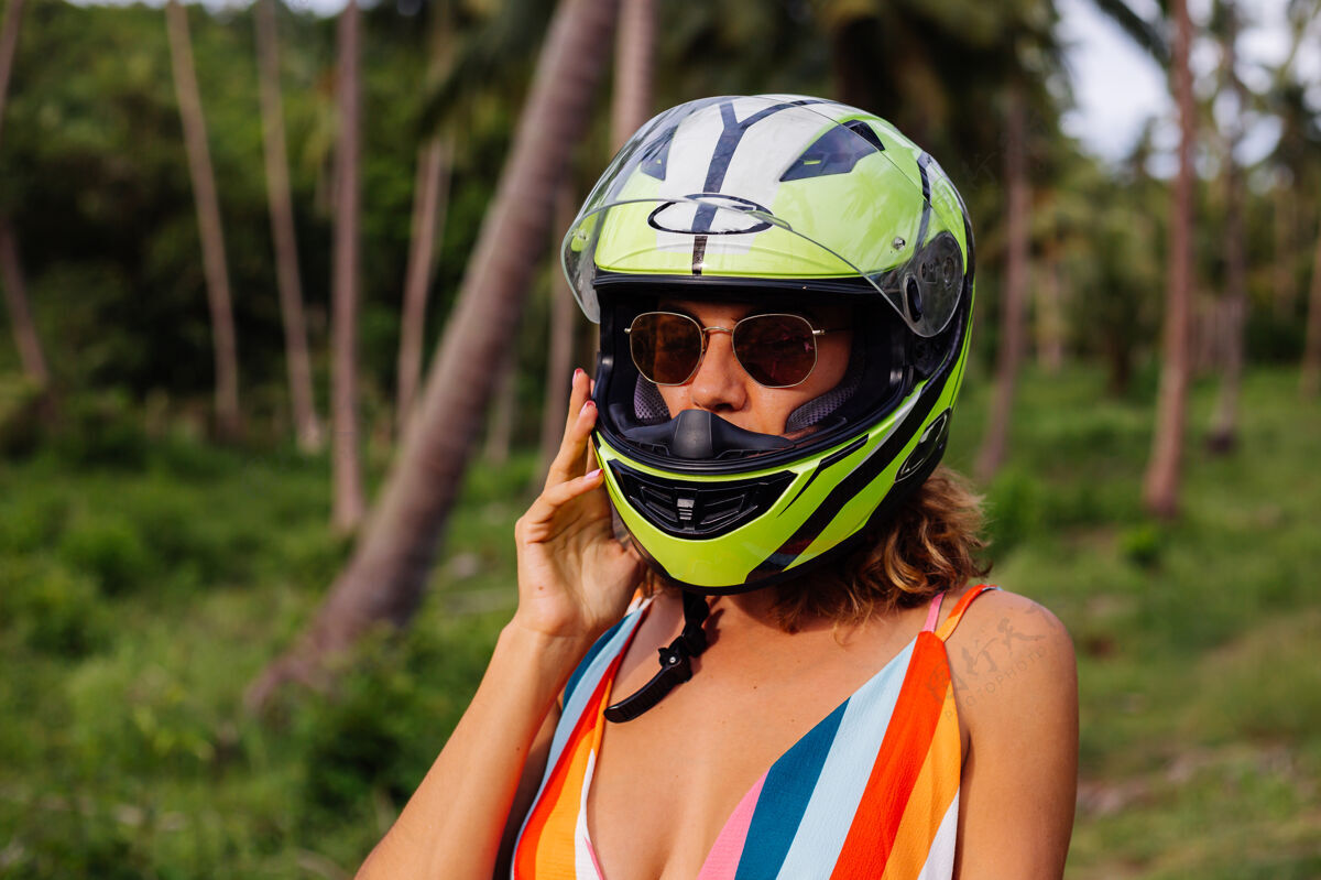热带在棕榈树下热带田野的丛林中 穿着黄绿色摩托车头盔和彩色浅夏装的美丽骑手妇女的肖像年轻度假热带