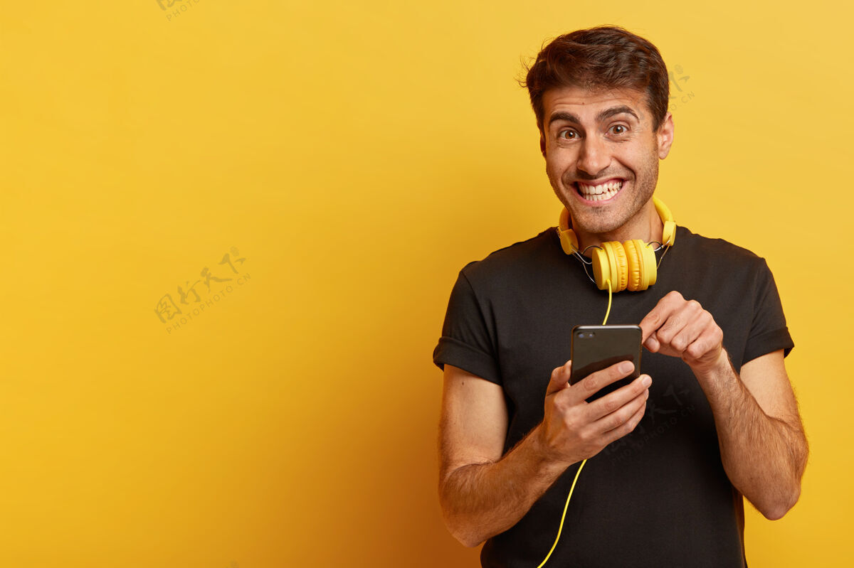 耳机高兴的欧洲人指着智能手机屏幕 戴着黄色耳机 穿着休闲的黑色t恤牙齿T恤表情