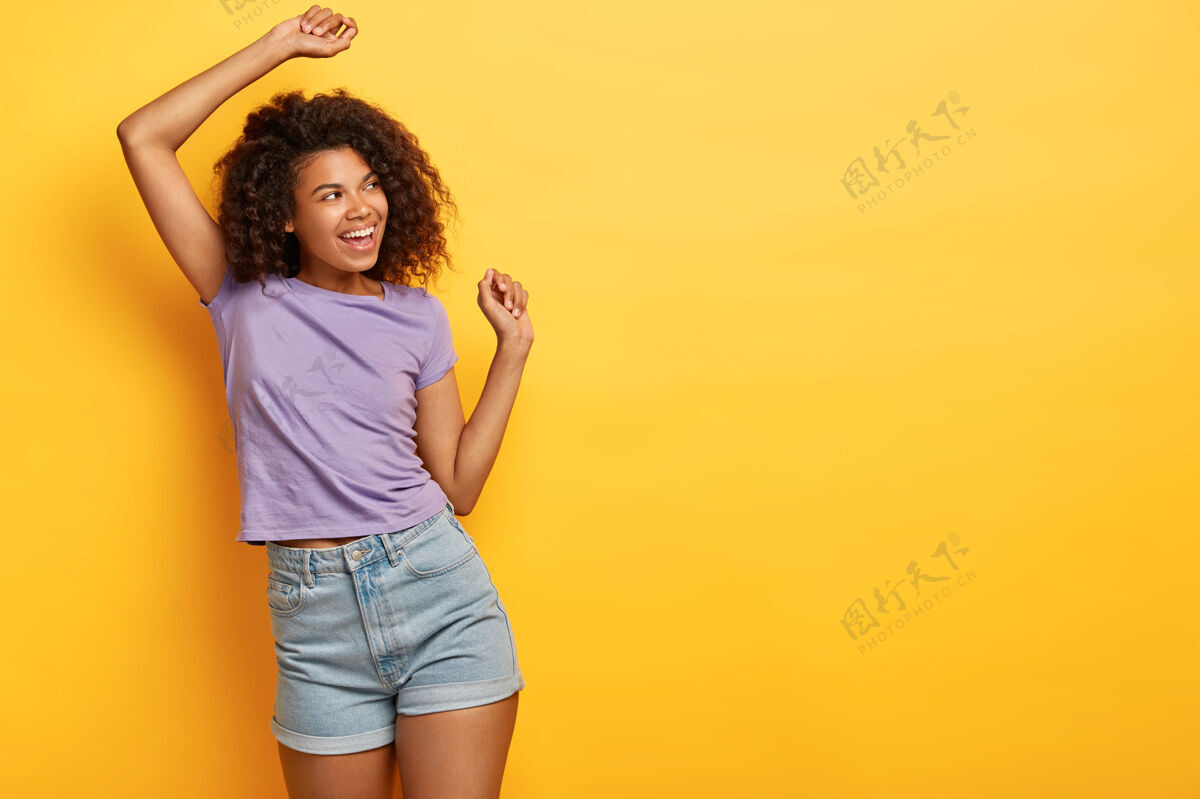 自由空间好幽默的非洲裔美国苗条女孩在黄色背景下跳舞 看起来很开心 穿着休闲的紫色t恤和牛仔短裤满足精力充沛牛仔