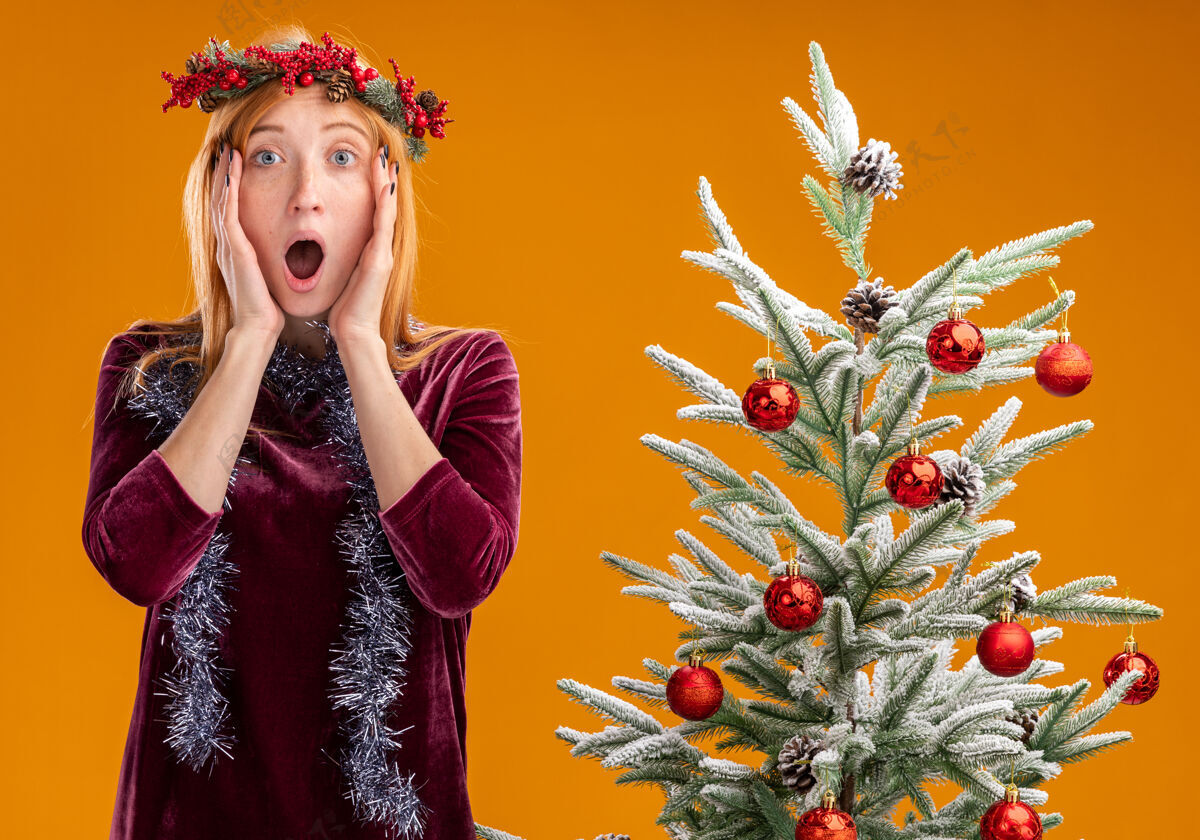 脖子站在圣诞树旁的年轻漂亮女孩 穿着红色的裙子 脖子上戴着花环 双手放在脸颊上 橙色背景下孤立着花环站着脸颊