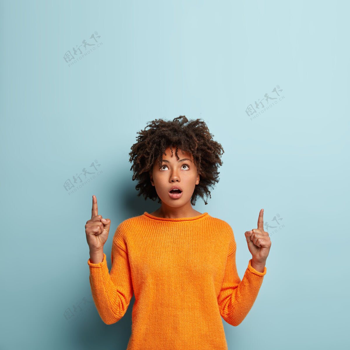 休闲惊艳美丽的黑人女子的室内镜头向上指 向上指方向 穿着橙色的套头衫 隔着蓝色的墙壁深肤色的女性促销对象情绪化印象深刻非裔美国人