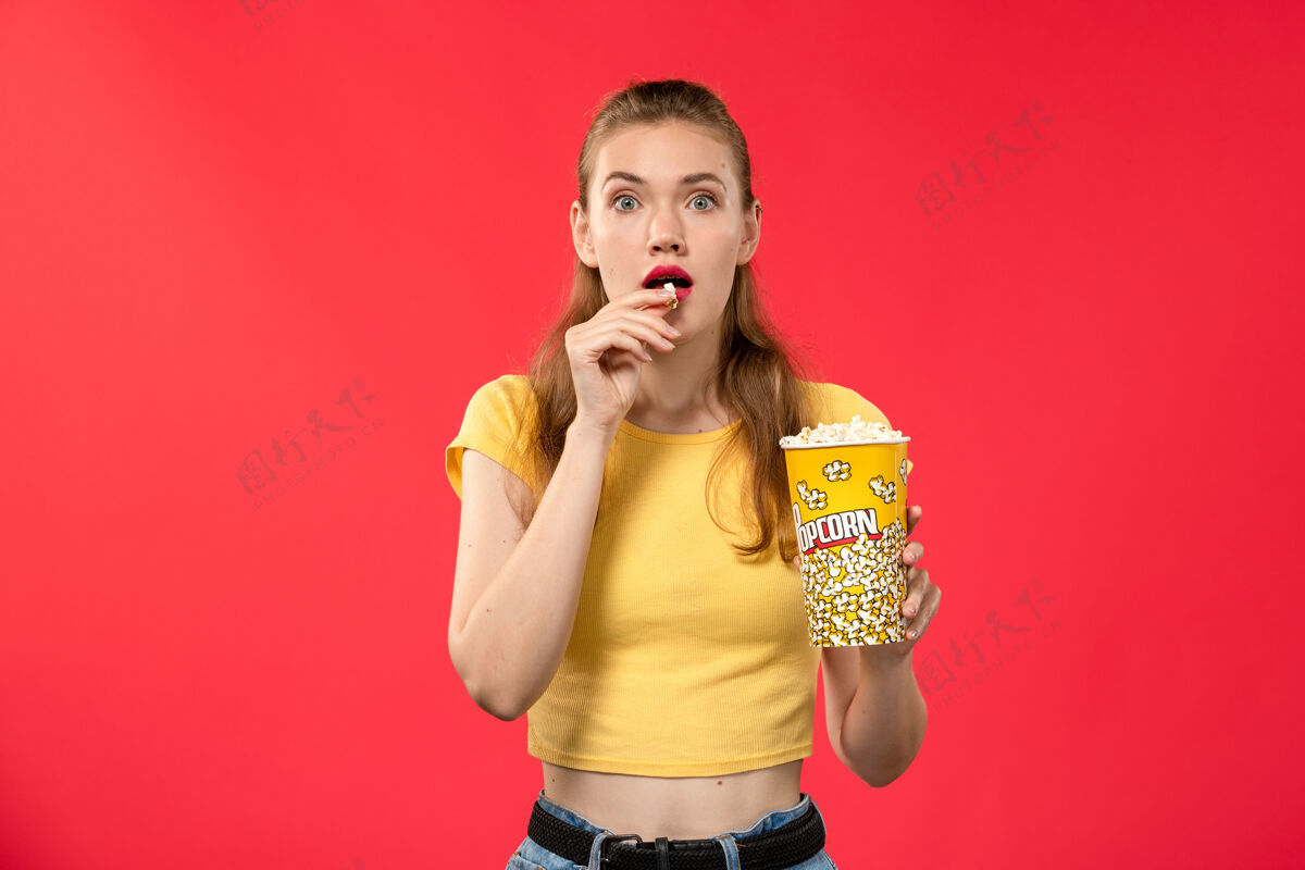 电影正面图年轻女性在电影院拿着爆米花包在浅红墙电影院看女性趣味电影灯光艺人爆米花