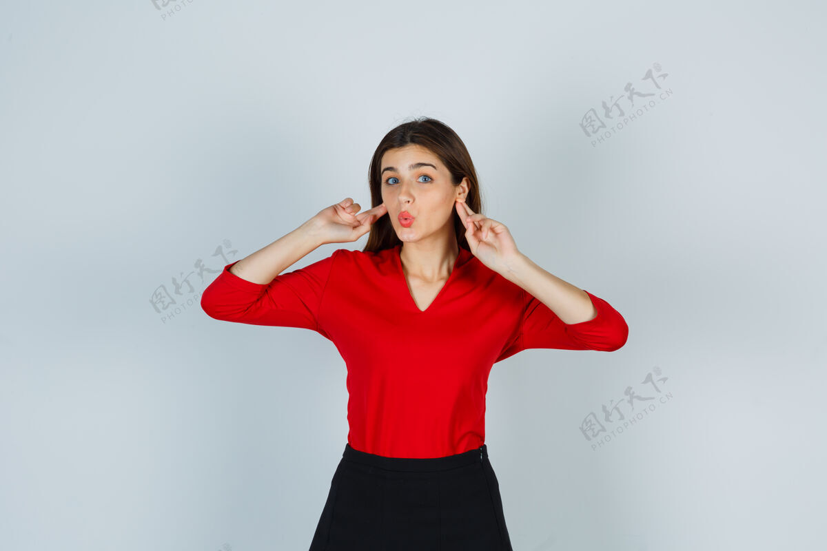女人穿着红衬衫的年轻女士 裙子把手指放在耳朵后面 看起来很滑稽衬衫情绪化裙子