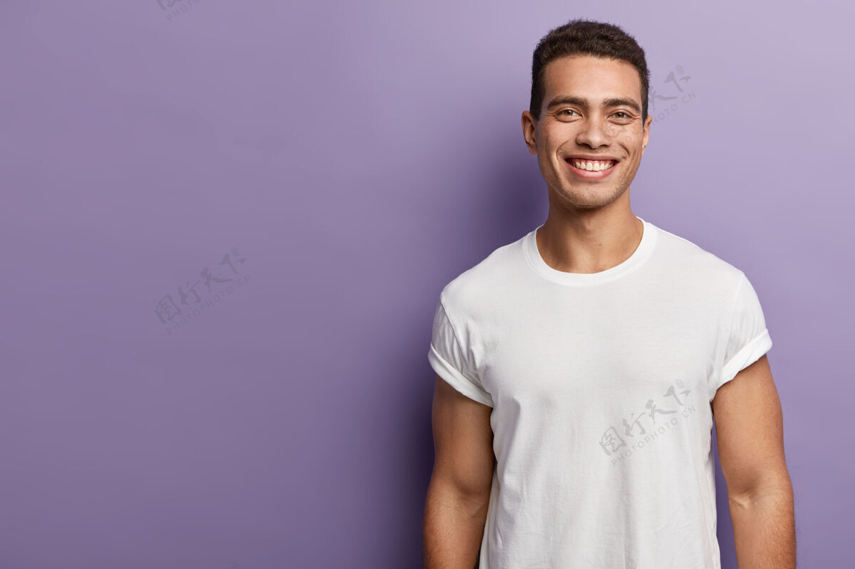 成人帅气开朗的年轻运动员有着运动的身体 肌肉发达的手臂 穿着白色的模拟t恤 深色短发 迷人的微笑 站在紫色的墙上 空白的复制空间旁边站立年轻外观