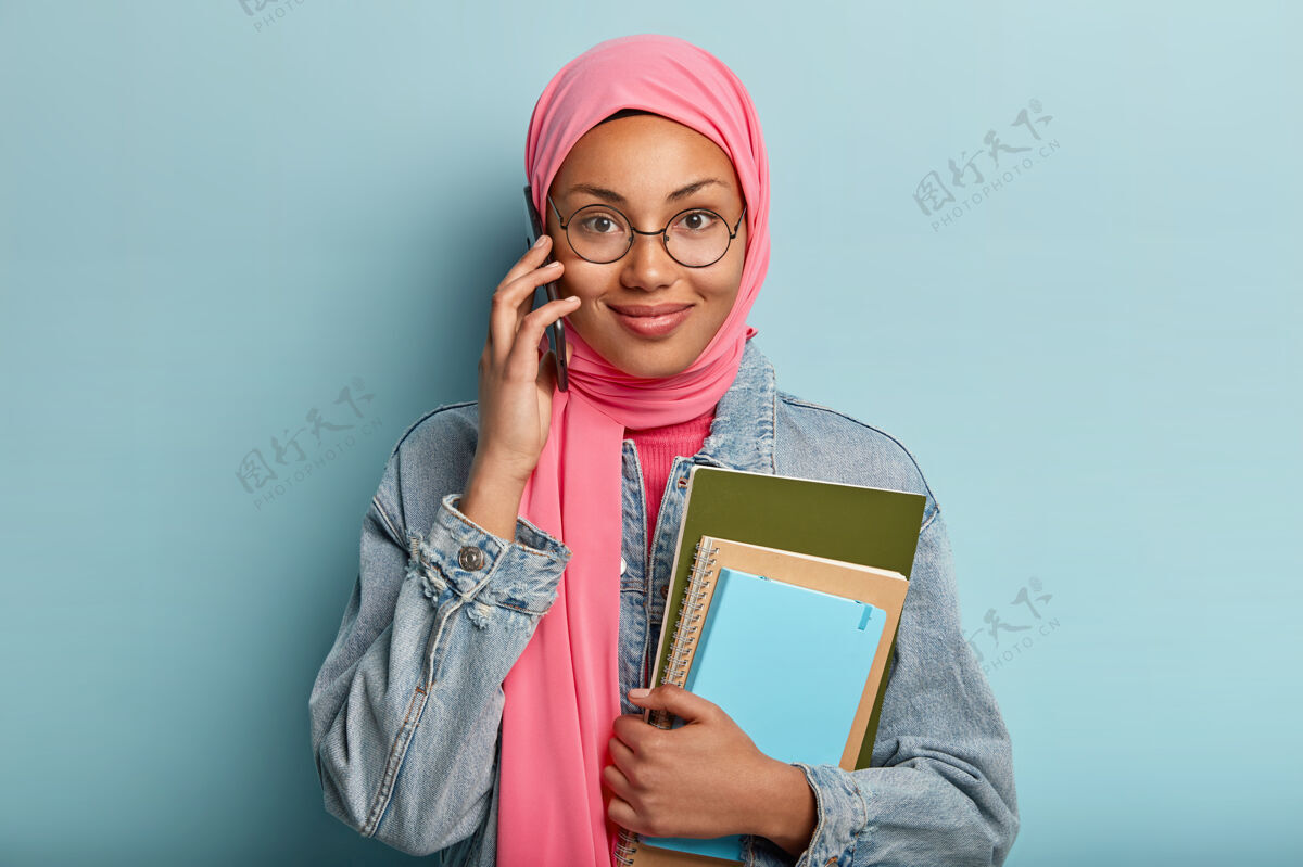 穆斯林满意的女性用手机聊天 闲暇时间闲聊 心满意足 拿着笔记本 穿着时髦的牛仔服 头上蒙着粉色的面纱 隔着蓝色的墙满意谈话眼镜