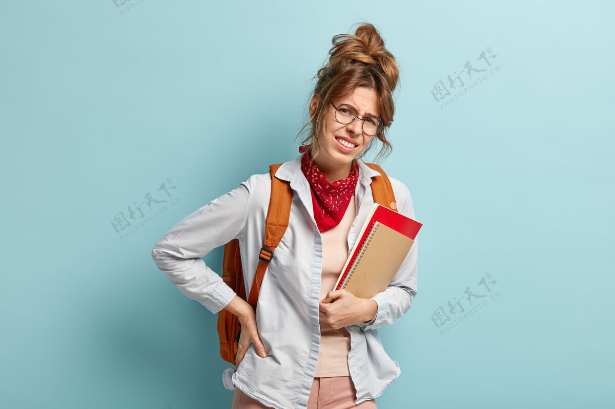 生病不快乐的女学生背着沉重的书包 背痛 拿着记事本 摸着腰 隔着蓝色的墙沮丧的青少年有健康问题时尚笔记本身体