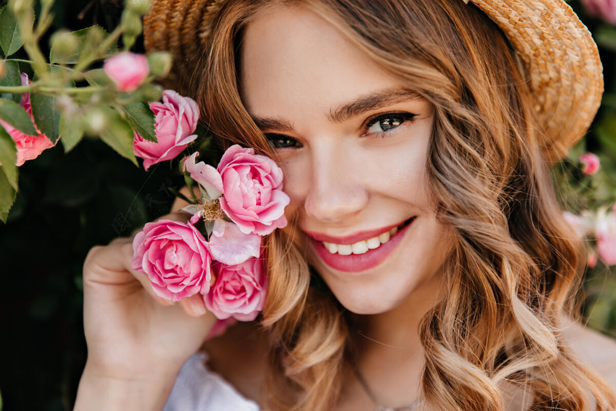 表情迷人女孩的特写肖像 闪亮的眼睛 与花摆姿势壮观的金发女子戴着帽子 手持粉红玫瑰 微笑着明亮女人树