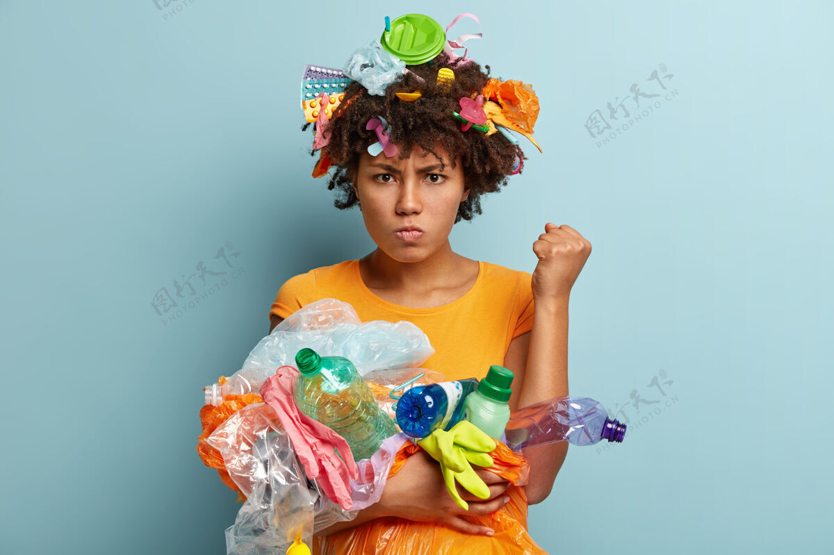 垃圾恼怒的黑人妇女的形象举起握紧的拳头 要求环保 有暴躁的面部表情 携带塑料垃圾 使用回收物品 站在蓝色的墙上环境保存不满