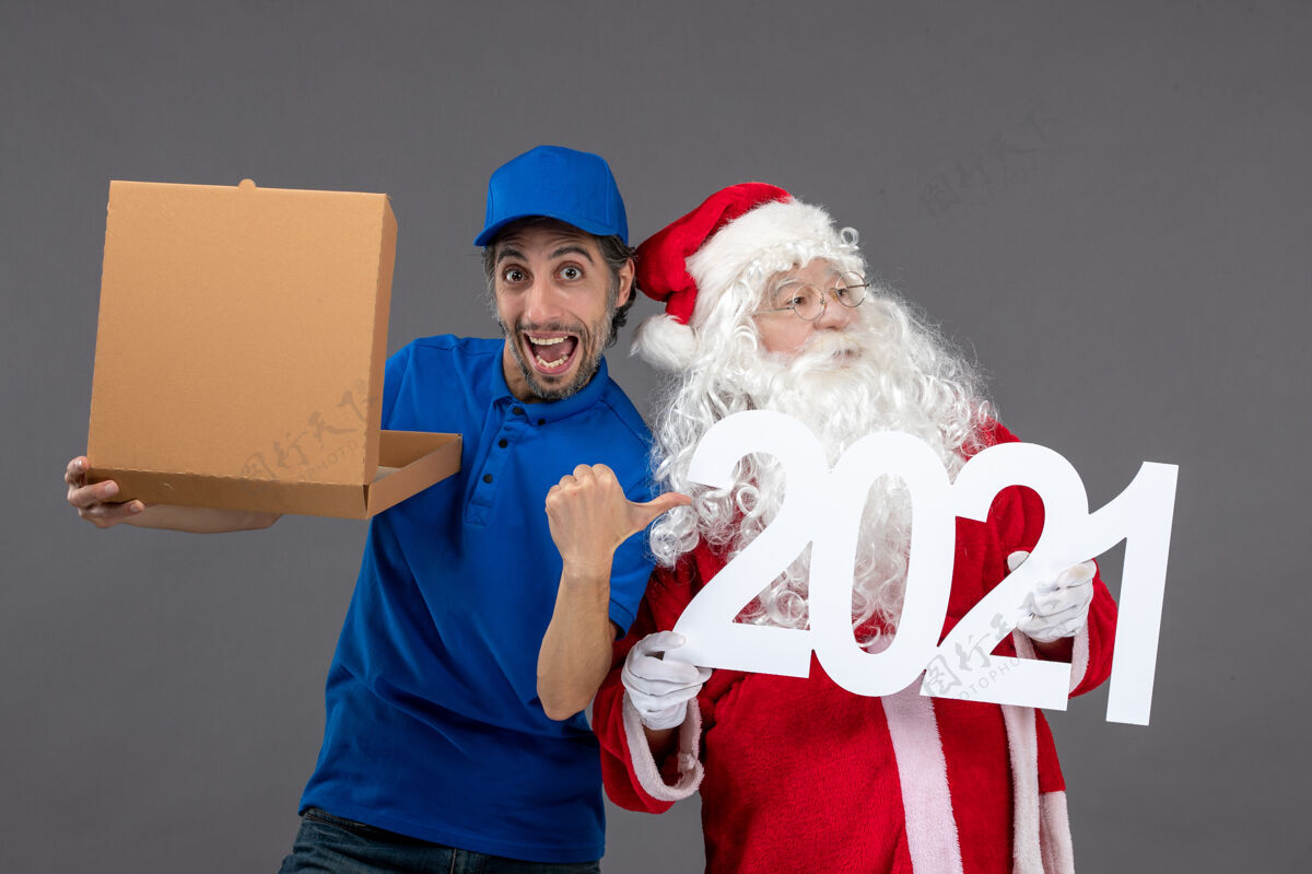帽子圣诞老人的正面图 男信使拿着食物盒 站在灰色的墙上服装正面圣诞老人