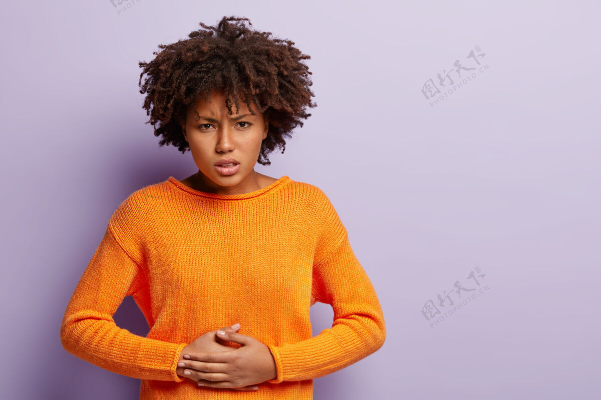 女人不满意的黑皮肤女性手放在肚子上 忍受疼痛 月经来潮 穿着橙色套头衫 卷发 隔离在紫色墙壁上 为你的广告腾出空间卷发腹部胃痛