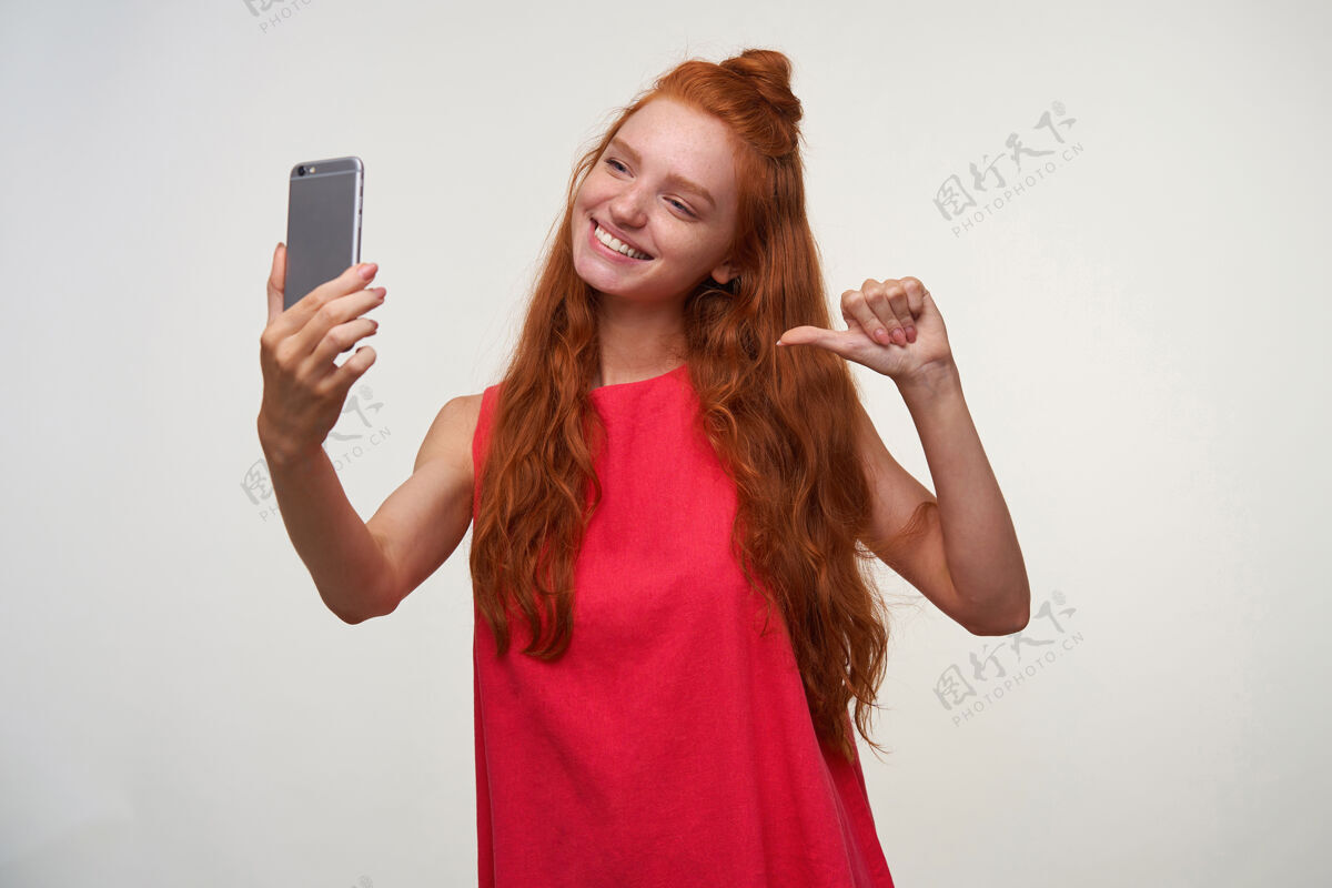 手机快乐的年轻readhead女性 头发打结 穿着休闲装 在白色背景上摆姿势 手里拿着手机 一边用拇指指着自己 一边自拍积极发型自拍