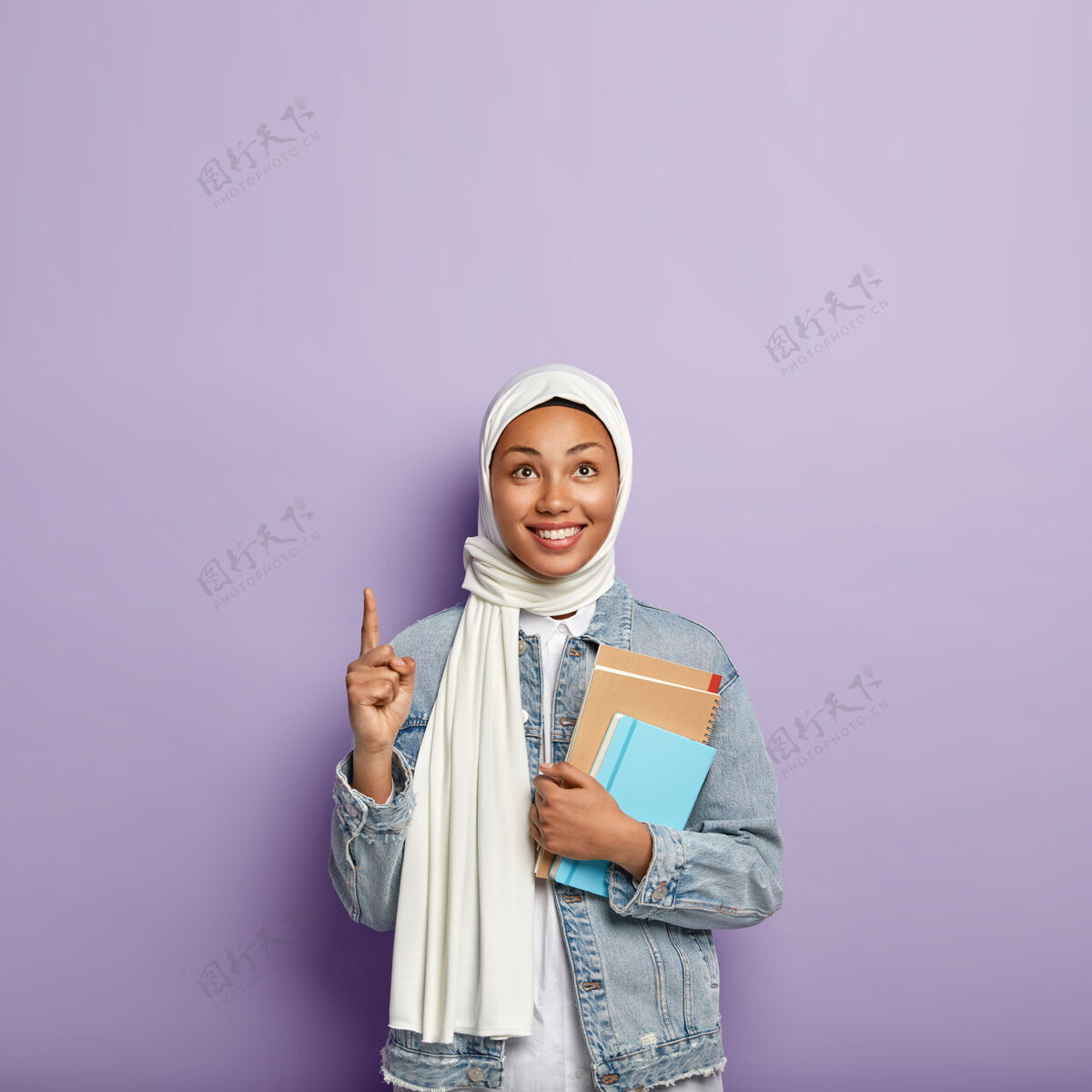 笔记本高兴的宗教妇女的孤立镜头用面纱盖住头 拿着记事本 用食指指着上面 高兴地笑着 站在紫色的墙壁上 空白处等待晋升伊斯兰女孩学习头巾个人表示