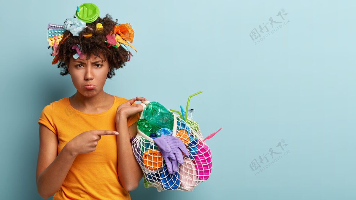 沮丧不满的黑皮肤女模特 捡起垃圾 不满地指着塑料垃圾 做志愿者 保护环境 站在蓝色的墙上 为你的文字腾出空间环保生态阴郁