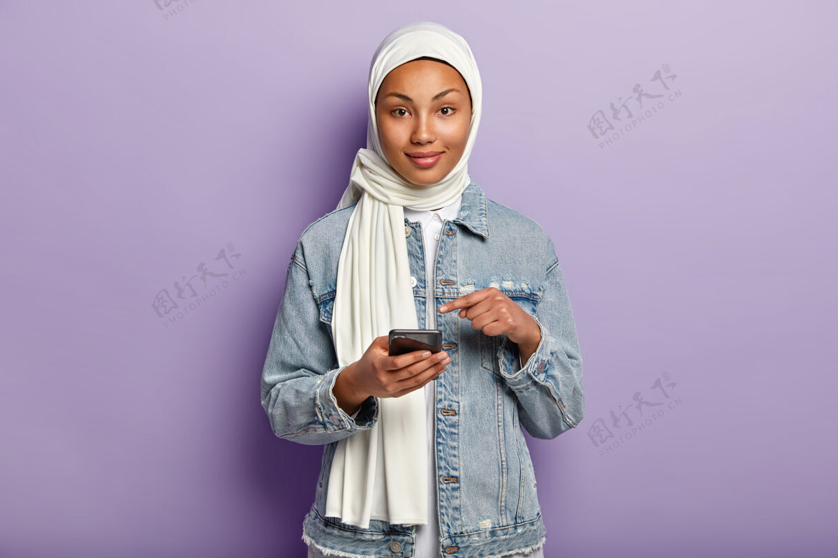 手机黑皮肤的快乐混血女孤立镜头 追随穆斯林宗教 指着手机屏幕 要求在网站上看网络新闻 孤立在紫色的墙上头巾短信手机