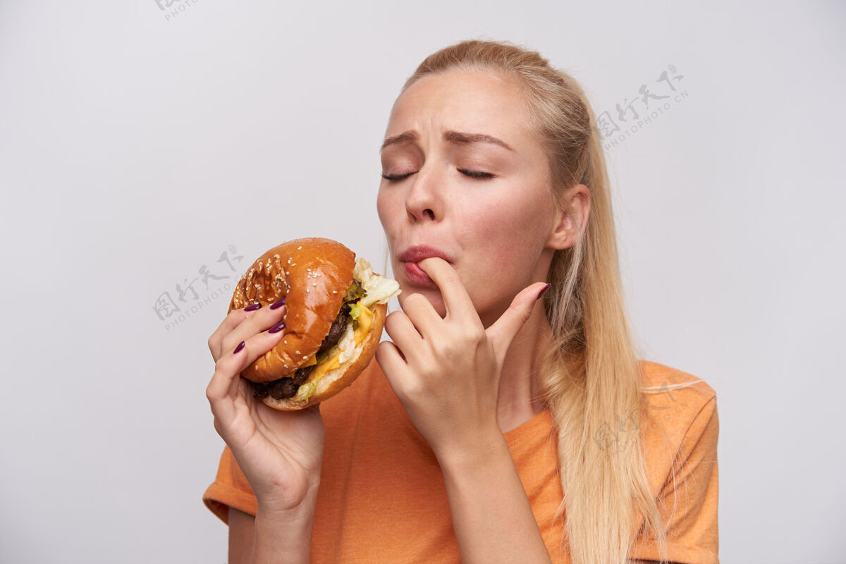 20多岁年轻而满意的金发女郎的肖像 随意的发型 非常愉快地品尝着她的新鲜汉堡包 紧闭双眼 站在白色的背景下室内饮食不健康