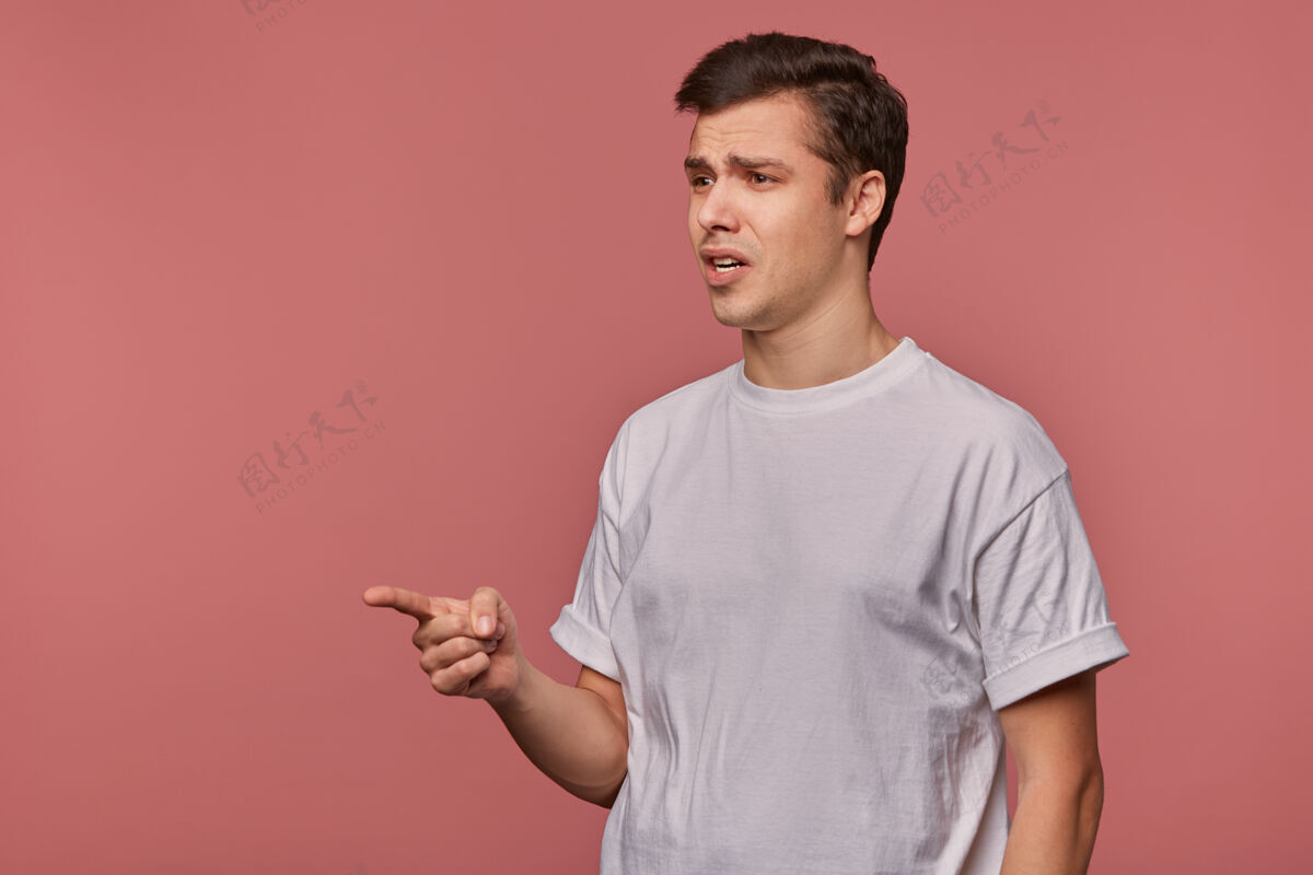 20多岁特写镜头：一个黑发的年轻人抬起食指指着一边 穿着休闲服在粉色背景上摆姿势 愁眉苦脸黑发男性深色