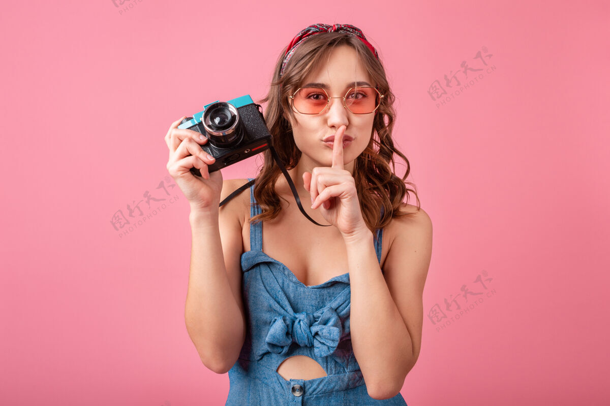 相机迷人的微笑女人展示沉默的手指标志在老式相机上拍照穿着牛仔裙隔离在粉红色背景上粉色连衣裙表情